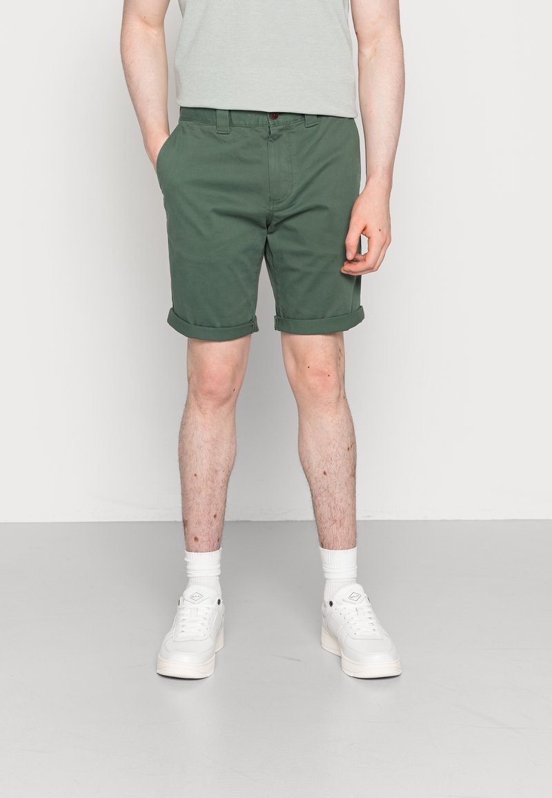Homme portant un short vert retroussé, des baskets blanches et des chaussettes blanches, debout, une main dans la poche, sur un fond clair uni.