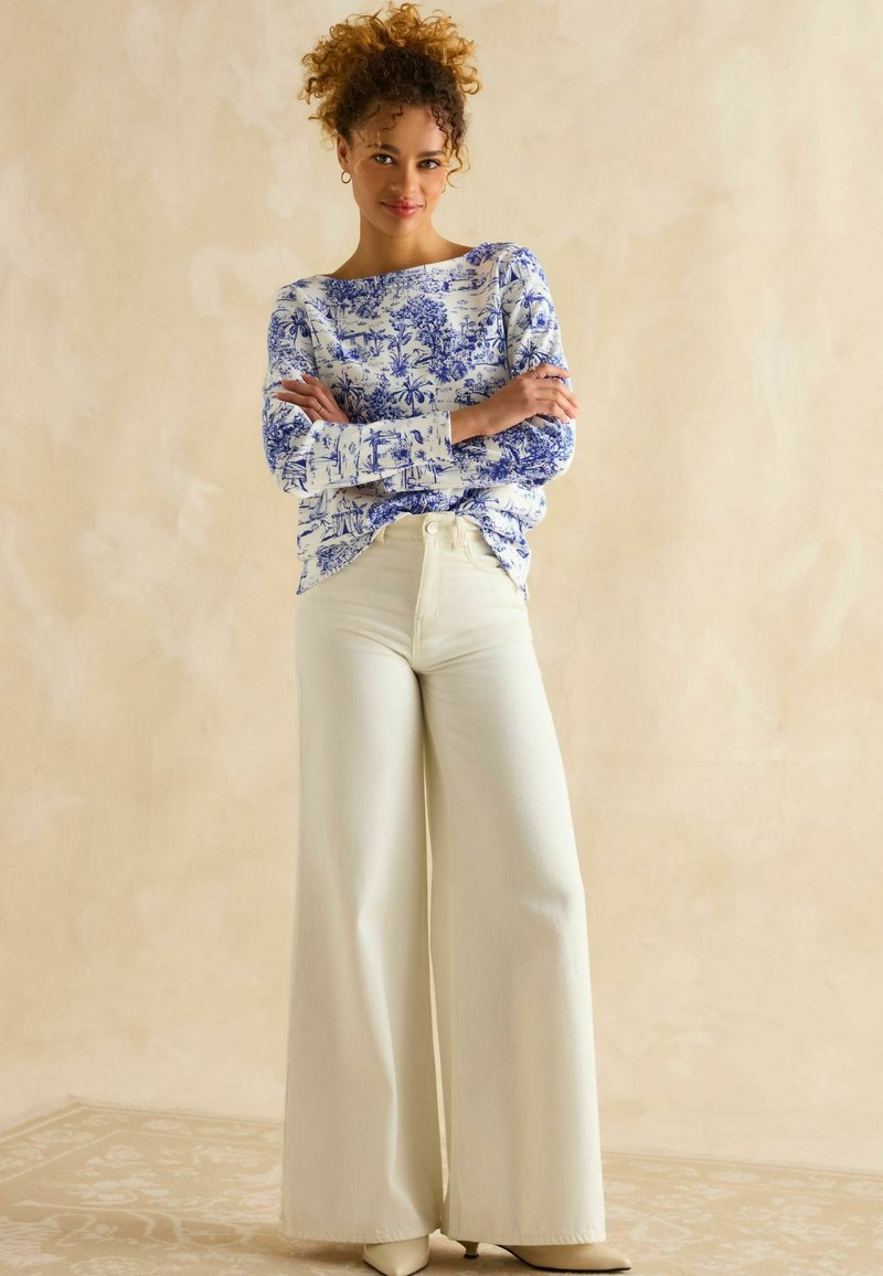 Langärmliges weißes Hemd mit blauer Blumenmuster auf den Ärmeln, kombiniert mit weißen, weit geschnittenen Hosen. Model trägt beigefarbene spitze Pumps.