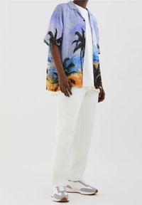 Hombre vestido con pantalones blancos, camisa blanca y una camisa de botones de manga corta con estampado de palmeras en azul, naranja y negro, complementada con zapatillas grises y blancas.