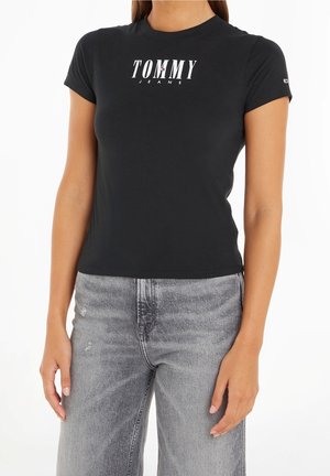 Kvinde iført en sort Tommy Jeans T-shirt og højttaljede, falmede grå jeans, stående med arme afslappet ned langs siderne.
