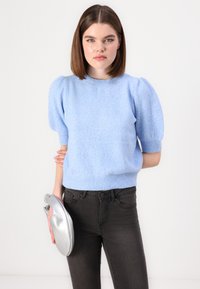 Suéter de punto azul claro con mangas abullonadas, cuello redondo y dobladillo acanalado. Combinado con jeans oscuros y un bolso de mano plateado.