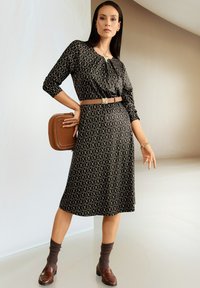 Frau in einem schwarzen gemusterten Midikleid mit einem braunen Gürtel und einer passenden quadratischen Handtasche, kombiniert mit braunen Socken und Slippern, steht drinnen.