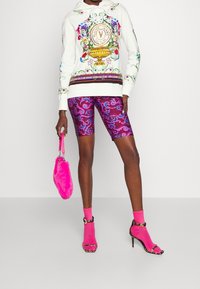 Mujer vistiendo un sudadera blanca con estampado floral, pantalones cortos de bicicleta morados con estampado, calcetines rosa neón con sandalias negras de tacón alto, sosteniendo un bolso de pelo rosa.
