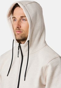 Felpa con cappuccio beige chiaro, dotata di zip full e lacci neri. Texture morbida e vestibilità casual, con un'etichetta logo ricamata.