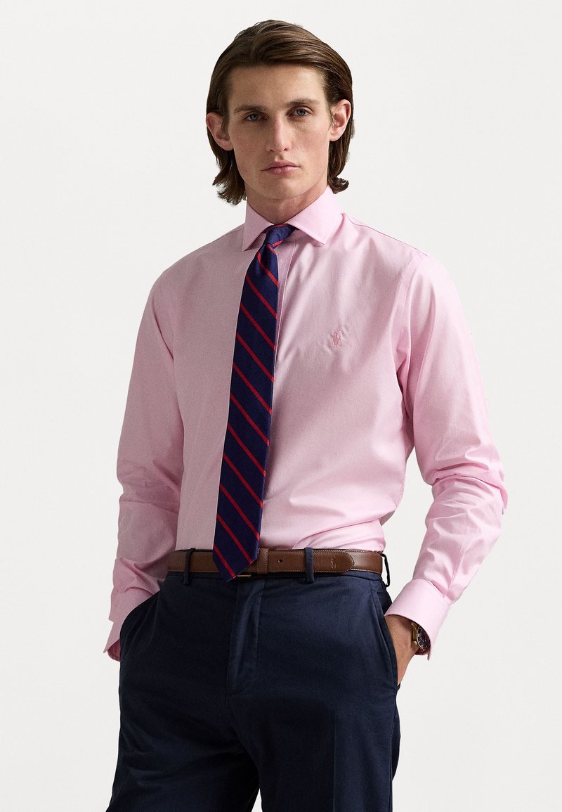 Cămașă roz cu mânecă lungă, asortată cu o cravată bleumarin cu dungi roșii. Cureaua maro și pantalonii bleumarin închis completează ținuta. Logo-ul subtil brodat pe cămașă.