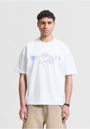 Giovane uomo con capelli corti e ricci che indossa una T-shirt bianca oversize con scritta colorata "Paternity Atelier" e pantaloni beige su uno sfondo semplice.