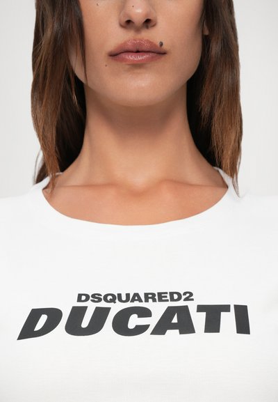 Ženska v belem srajcu z black "DSQUARED2 DUCATI" napisom, ki prikazuje ustnice z obrazno madežem in dolge rjave lase.