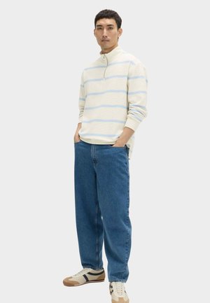 Uomo che indossa un maglione beige e azzurro a righe con cerniera a quarto, jeans blu e sneakers beige con dettagli blu navy e marroni, in piedi con le mani nelle tasche.