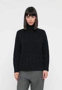 Maglione a collo alto lavorato a maglia nera con motivo a trecce, polsini e orlo a coste, indossato su pantaloni grigi a righe.