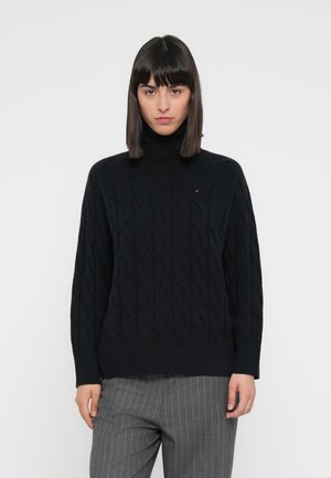 Maglione a collo alto lavorato a maglia nera con motivo a trecce, polsini e orlo a coste, indossato su pantaloni grigi a righe.