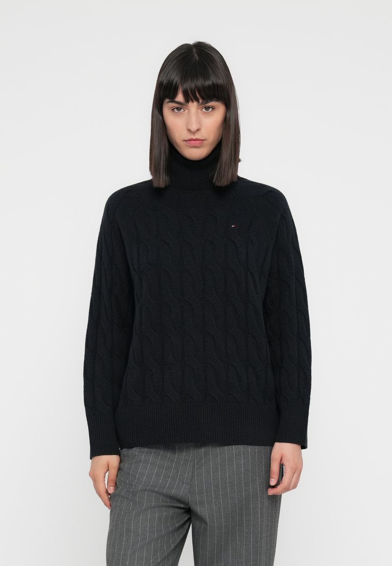 Maglione a collo alto lavorato a maglia nera con motivo a trecce, polsini e orlo a coste, indossato su pantaloni grigi a righe.