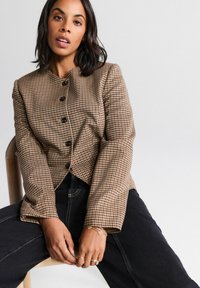 Brown and beige checkered blazer with a round neckline, black buttons, and flared sleeves, paired with dark denim jeans.