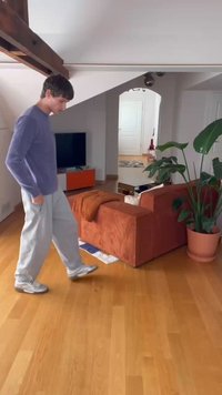 Pantaloni de trening gri deschis și o cămașă lungă mov purtate de o persoană care merge pe podeaua din lemn, aproape de o canapea portocalie din catifea și o plantă.
