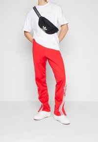 Osoba stojí oblečená v bílých teniskách, jasně červených teplácích Adidas s bílými pruhy a logy, bílém tričku a černé crossbody tašce Adidas.