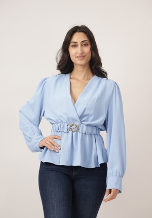 Vrouw met lang donker haar die een lichtblauw blouse met ceintuur en donkere spijkerbroek draagt, staande met één hand op haar heup tegen een neutrale achtergrond.