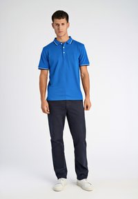 Niebieska koszulka polo z krótkim rękawem, z kontrastowym białym kołnierzykiem i akcentami na rękawach, w zestawie z ciemnonawieszymi spodniami i białymi trampkami.