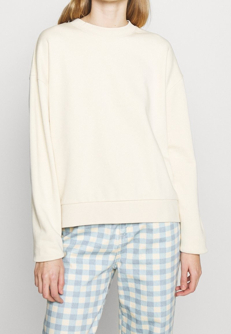 Felpa beige chiaro con scollatura rotonda, vestibilità rilassata e maniche lunghe. Tessuto morbido e orlo a coste, abbinata a pantaloni a scacchi bianco e blu.