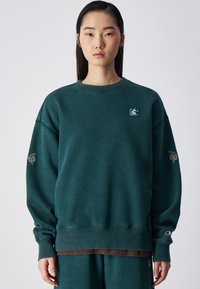 Zielona bluza z luźnym krojem, prążkowanym dekoltem w kształcie Crew i haftowanym logo. Długie rękawy mają ozdobne szwy i akcenty.