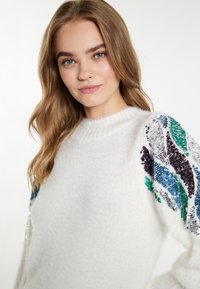 Witte trui met een donzige textuur, voorzien van veelkleurige paillettenpatronen op de mouwen in tinten blauw, groen, paars en zilver.