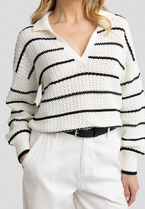 Femme portant un pull en maille blanc avec des rayures horizontales noires et un col, associé à un pantalon blanc et une ceinture noire.
