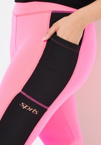 Rosa och svarta sportleggings med en sidficka, kontrasterande paneler och en logotyp "sport" på den svarta delen. Slät textur.