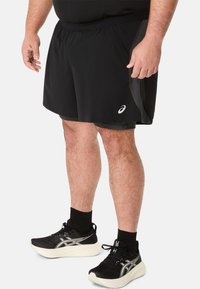 Zwarte sportshorts met een gladde textuur, voorzien van een zijzak en een elastische tailleband. Combinatie met zwarte hardloopschoenen en sokken.