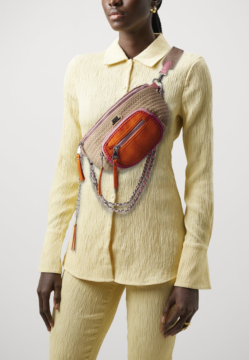 Sac banane en matériau tissé beige avec des accents orange et rose, détails en chaîne argentée et poches zippées. Porté par-dessus un ensemble texturé jaune.