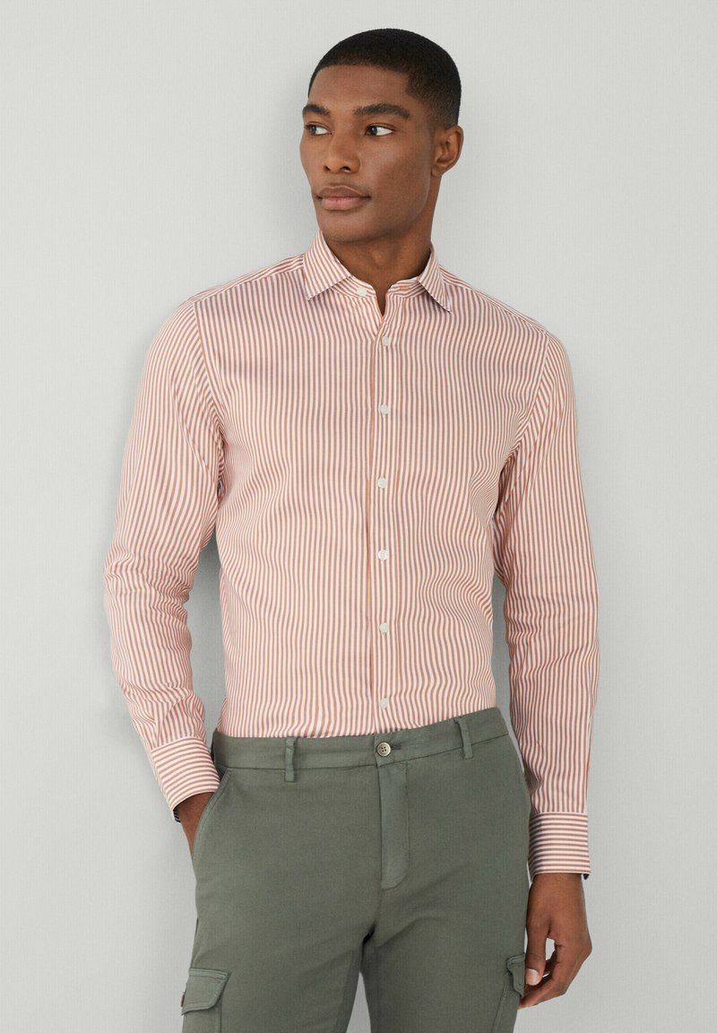 Une chemise à boutons avec des rayures verticales rouges et blanches, à manches longues et col. Associée à un pantalon vert, mettant en valeur une coupe ajustée.