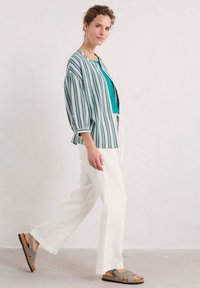 Gestreepte lichtgewicht jas in groen en wit, teal top, witte wijde broek en grijze sandalen met kurken zool en gekruiste banden.