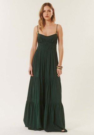 Jeune femme aux cheveux ondulés portant une robe longue sans manches vert foncé avec de fines bretelles et une jupe à volants, debout devant un fond uni.
