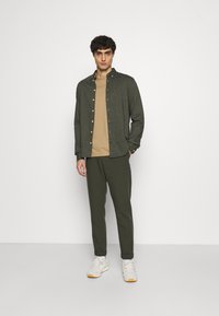 Grüne Button-Down-Hemd über einem beigen T-Shirt, kombiniert mit dunkelgrünen Hosen und hellen Sneakers. Das Hemd hat einen strukturierten Stoff mit sichtbaren Knöpfen.