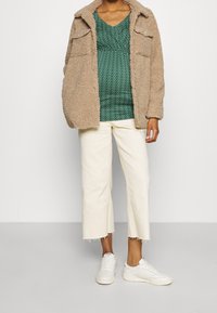 Béžová fleecová bunda, zelený vzorovaný top, bílé zkrácené kalhoty a bílé tenisky. Bunda má dvě přední kapsy a límeček.