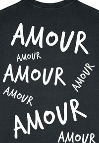 Juodas marškinėlis su žodžiu "AMOUR" baltomis raidėmis, įvairių dydžių ir orientacijų, minkšta tekstūra ir apvaliu kaklu.