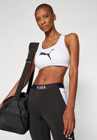 Hvit sports-bh med svart Puma-logo, sammen med svart og hvit leggings med en Puma livrem, holder en svart treningspose.