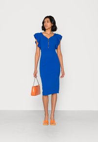 Vestido azul justo com mangas rendadas e fecho éclair frontal. A modelo segura uma mala em padrão de crocodilo laranja e usa sandálias de salto laranja.