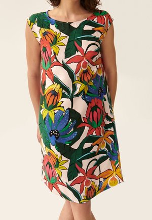 Ærmeløs kjole med livligt tropisk blomstermønster i rød, gul, blå og grøn på en lys baggrund, båret af en stående person.