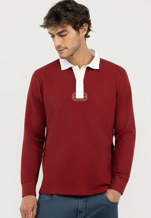 Hombre con camisa polo roja de manga larga con cuello blanco y tapeta con botones, mirando hacia abajo, una mano en el bolsillo del pantalón azul.
