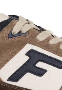 Primo piano di una sneaker marrone e bianca con lacci beige e una lettera "F" blu scuro cucita sul pannello laterale.