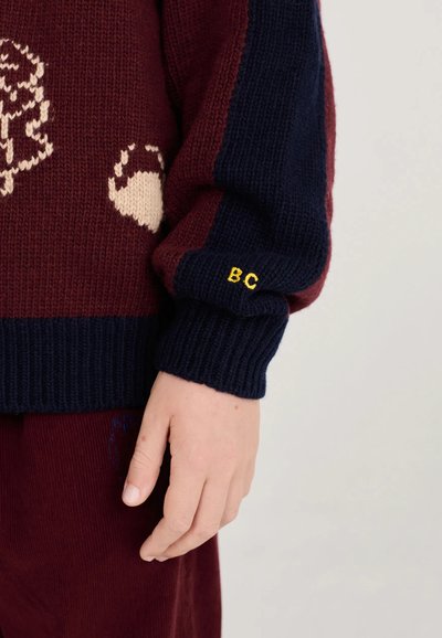 Gros plan d'une main portant un pull en laine à rayures bordeaux et bleu marine avec les initiales BC brodées en jaune sur le poignet.