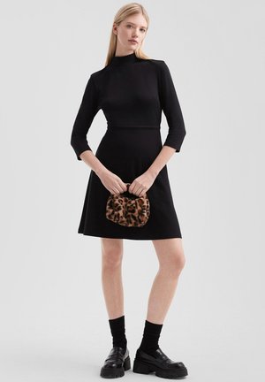 Robe noire à manches trois-quarts, buste ajusté, jupe évasée ; accessoirisée avec un sac à main en fausse fourrure léopard et des chaussures noires à semelles épaisses.