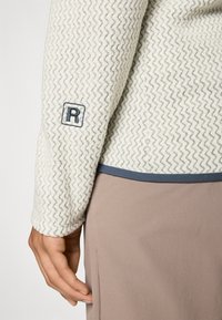 Nahaufnahme eines Arms in einer strukturierten, weißen Jacke mit Zickzackmuster und dunklem "R"-Logo auf dem Ärmel, kombiniert mit hellbraunen Hosen.