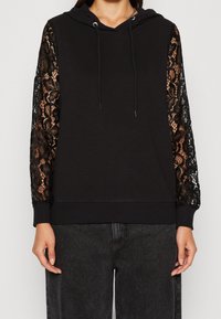 Sudadera negra con mangas largas de encaje, que presenta una capucha con cordones y un dobladillo acanalado. El tejido principal es suave, en contraste con la delicada textura del encaje.