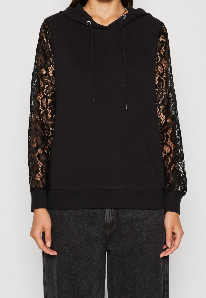 Sudadera negra con mangas largas de encaje, que presenta una capucha con cordones y un dobladillo acanalado. El tejido principal es suave, en contraste con la delicada textura del encaje.