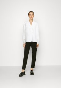 Weiße, strukturierte Bluse mit V-Ausschnitt und Knopfleiste, kombiniert mit schwarzen, taillierten Hosen und glänzenden schwarzen Schuhen.