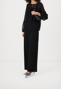 Femme portant un blouse noire transparente à manches longues, un pantalon noir à jambes larges, des escarpins argentés pointus, et portant un sac à bandoulière noir clouté.