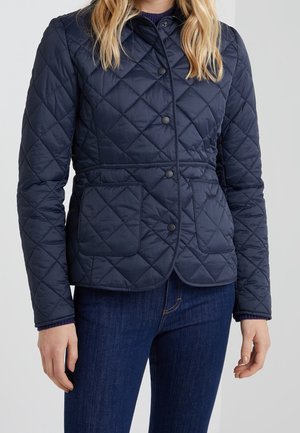 Femme portant une veste matelassée bleu marine avec des boutons-pression et deux poches avant, associée à un jean bleu foncé.