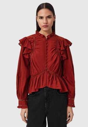 Czerwona bluzka z falbanami na ramionach i wysokim kołnierzem, z zapięciem na guziki i elastycznym pasem. Połączona z czarnymi spodniami.