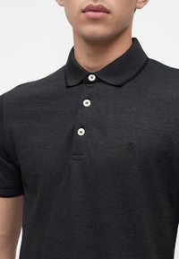 Homme portant un polo gris foncé avec un col texturé et trois boutons blancs sur la patte de boutonnage, sans autres détails visibles.
