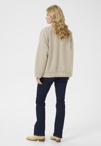 Pull oversized beige avec taille élastique, associé à un jean flare bleu foncé et des bottines beiges. Texture lisse, design minimal.