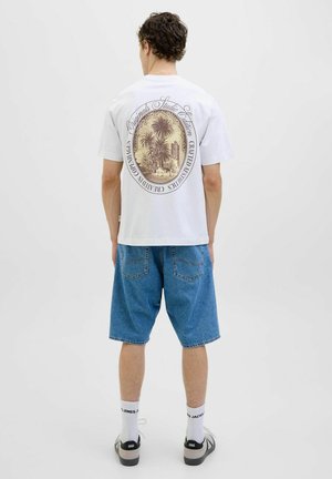 Hvid t-shirt med et stort, retro-stil grafik med palmer og tekst på ryggen. Båret med blå denim shorts og sorte sko.